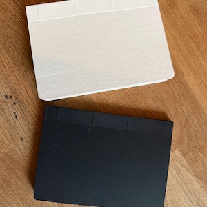 Puede incluir: Dos cuadernos rectangulares, uno con cubierta blanca sólida y otro con cubierta negra sólida. Ambos cuadernos presentan una encuadernación cosida a lo largo del borde superior con segmentos de hilo visibles, orientados horizontalmente.