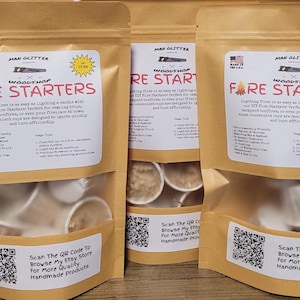 May include: Three brown paper bags with a white label that says "Woodshop Fare Starters" and a QR code. The bags contain small white fire starters. The bags are on a wooden surface.