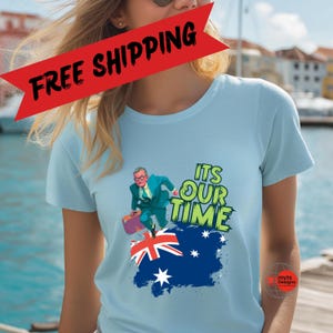 May include: Light blue t-shirt featuring a cartoon of a man in a suit running with a suitcase over an Australian flag. The text "ITS OUR TIME" is in green and white. The shirt has a round neck and short sleeves.