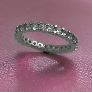 Puede incluir: Un anillo de eternidad plateado con una fila continua de piedras preciosas redondas y transparentes. El anillo se muestra sobre una superficie rosa, resaltando el brillo de las piedras. La banda tiene un diseño detallado.