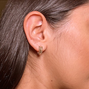 May include: Gold-coloured hoop earrings with a leaf design. The earrings are small and hug the earlobe. The design features a series of small, detailed leaves arranged around the hoop. The earrings are shown on a person's ear.