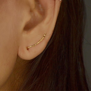 May include: A gold ear climber earring with a textured, curved design. The earring is worn on the ear, showcasing its delicate and minimalist style. This piece of jewellery adds a touch of elegance.