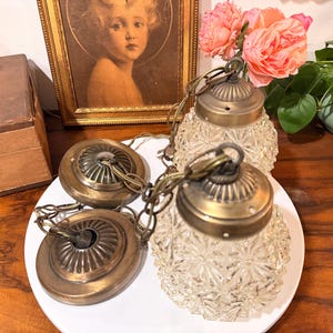 May include: Two pendant lights featuring clear, textured glass shades with a starburst pattern and antique brass-toned metal hardware. Each light includes a matching circular ceiling canopy and a linked chain for suspension, showcasing a classic design.