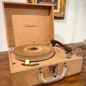 May include: A portable Silvertone record player in a tan, suitcase-style case. The interior lid features the brand name Silvertone and a handwritten name. It includes a circular turntable, a brown tone arm, a speaker grille, and a clear handle.
