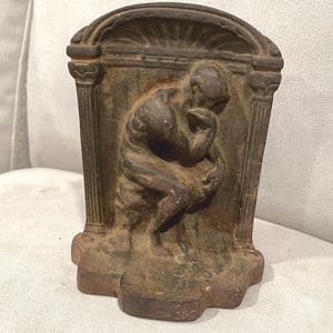 May include: A weathered, bronze-colored cast iron bookend featuring a sculpted figure in a seated, contemplative pose. The figure rests on a base with architectural columns and an arched top. The bookend is designed to support books on a shelf.