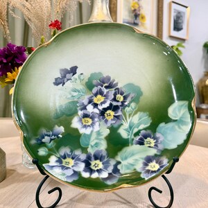 May include: A decorative, scalloped-edge plate with a green and white gradient background. The plate features a floral design of purple and yellow flowers with green leaves. The rim is trimmed with gold. The plate is displayed on a black metal stand.