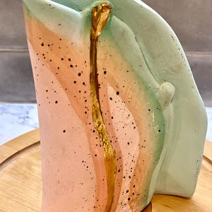 May include: A ceramic vase with a unique, abstract design. The vase is painted in shades of pink, green, and gold, with a gold stripe running down the center. The vase has a speckled texture and a curved shape.
