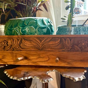 May include: A rectangular wooden wall rack featuring a horizontal panel with a carved floral and leaf pattern. The rack includes four evenly spaced wooden pegs extending outward from the lower section, showcasing a natural wood grain finish.