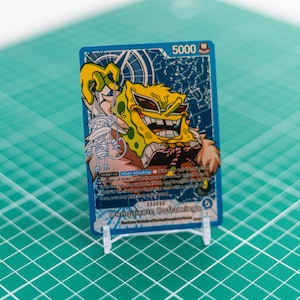 Könnte beinhalten: Blaue Sammelkarte mit einem gelben Cartoon-Charakter mit Sonnenbrille und breitem Grinsen. Die Karte, auf einem transparenten Kunststoffständer, hat eine Stärke von 5000. Der Hintergrund ist ein grünes Raster.