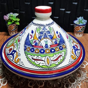 Könnte beinhalten: Ein dekorativer Keramik-Tajine-Topf mit konischem Deckel. Der weiße Topf hat aufwendige, handgemalte Designs in Blau, Grün, Gelb und Rot. Der Rand und der Deckel sind mit blauen und roten Bändern verziert. Ein traditionelles Kochgefäß.
