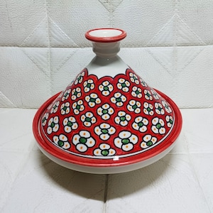 May include: A ceramic tagine pot with a red base and lid, decorated with a floral pattern in white, green, and yellow on a red backdrop. The pot features a conical lid and a circular base, ideal for Moroccan cuisine.