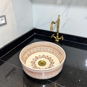 Puede incluir: Un lavabo decorativo de cerámica con un patrón geométrico marrón y blanco. El lavabo redondo está sobre una encimera negra. Un grifo de latón está montado detrás del lavabo.