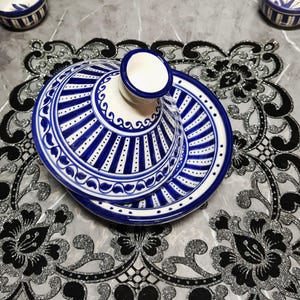 Puede incluir: Una olla de tagine de cerámica con base blanca y patrones geométricos azules. La tapa tiene un asa cónica. La olla se encuentra sobre un mantel individual floral decorativo negro y plateado. Se ven dos cuencos a juego en el fondo.