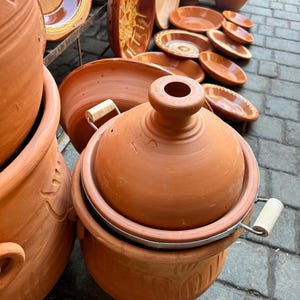 Könnte beinhalten: Eine Sammlung von Kochgeschirr aus Terrakotta, darunter ein Tajine-Topf mit konischem Deckel und Holzgriffen sowie verschiedene Servierschalen. Die Keramik hat einen warmen, rötlich-braunen Farbton, der auf traditionelle Handwerkskunst und kulinarische Verwendung hindeutet.