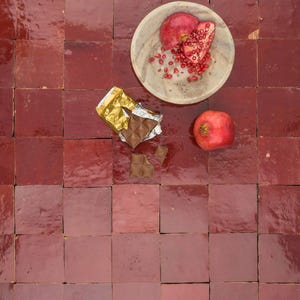 May include: A top-down view of a wooden bowl holding pomegranate halves and seeds, alongside a whole pomegranate and a partially unwrapped chocolate bar. These items are set on a square, red-tiled surface, offering a contrast of textures and colours. The tiles are approximately 15 cm x 15 cm.