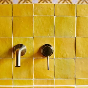 Pode incluir: Close-up de uma parede de banheiro com azulejos quadrados amarelos brilhantes e rejunte branco. Duas torneiras de níquel escovado são montadas na parede. A parte superior da parede tem uma borda de azulejo decorativa com um padrão repetitivo.