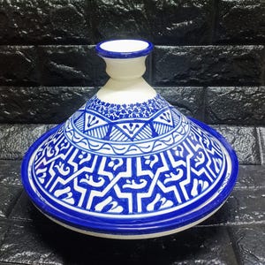 May include: A white and blue ceramic tagine pot with intricate geometric patterns. The lid and base are adorned with detailed blue designs on a white background, creating a striking contrast. The pot has a conical lid and a wide base.