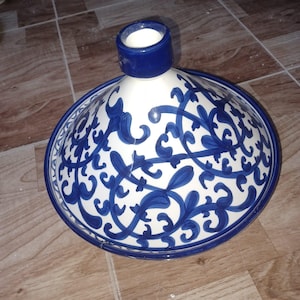Puede incluir: Una olla de tagine de cerámica blanca y azul con tapa cónica. La olla presenta un detallado patrón floral azul y un borde azul oscuro. La tapa tiene un pequeño asa cilíndrica. El tagine es un recipiente de cocina marroquí tradicional.