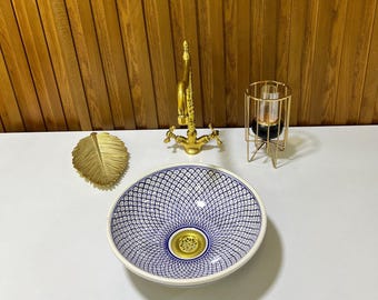May include: A white ceramic sink with a blue and white geometric pattern. The sink has a gold-coloured drain and is sitting on a white countertop.