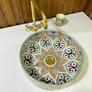 May include: A white ceramic sink with a green, yellow, and blue floral pattern. The sink has a round shape and a gold drain.