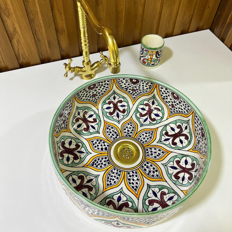 May include: A white ceramic sink with a green, yellow, and blue floral pattern. The sink has a round shape and a gold drain.