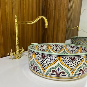 May include: A round ceramic sink with a colorful Moroccan-inspired design. The sink features a green, yellow, blue, and red pattern with intricate details. A gold faucet is mounted above the sink.