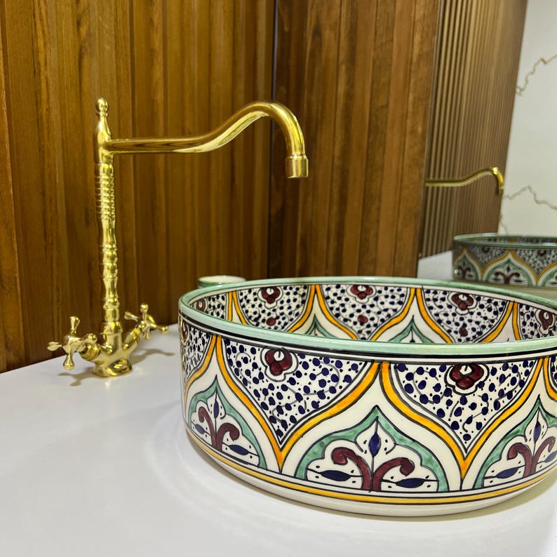 May include: A round ceramic sink with a colorful Moroccan-inspired design. The sink features a green, yellow, blue, and red pattern with intricate details. A gold faucet is mounted above the sink.