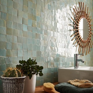 Pode incluir: Uma casa de banho com uma parede de azulejos quadrados turquesa. Um espelho redondo com uma moldura de bambu está acima de uma pia branca. Um cacto e outras plantas estão em uma superfície de madeira, junto com uma escova e uma toalha dobrada.
