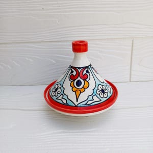 May include: A ceramic tagine pot with a conical lid and a round base. The pot is white with a red rim and lid top. The lid is decorated with a blue, red, and yellow floral pattern. The tagine is a traditional cooking vessel.