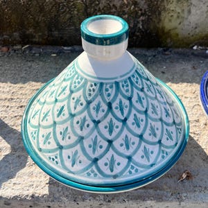 Puede incluir: Una olla de tagine de cerámica con tapa cónica y base. La olla es blanca con un patrón turquesa y azul. La tapa tiene un borde turquesa y una parte superior blanca. La base es turquesa. El patrón es un diseño repetido de hojas estilizadas.