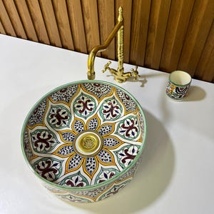 May include: A round ceramic sink with a colorful, intricate floral pattern in shades of green, yellow, red, and blue. The sink has a gold-colored drain and is set against a white background.