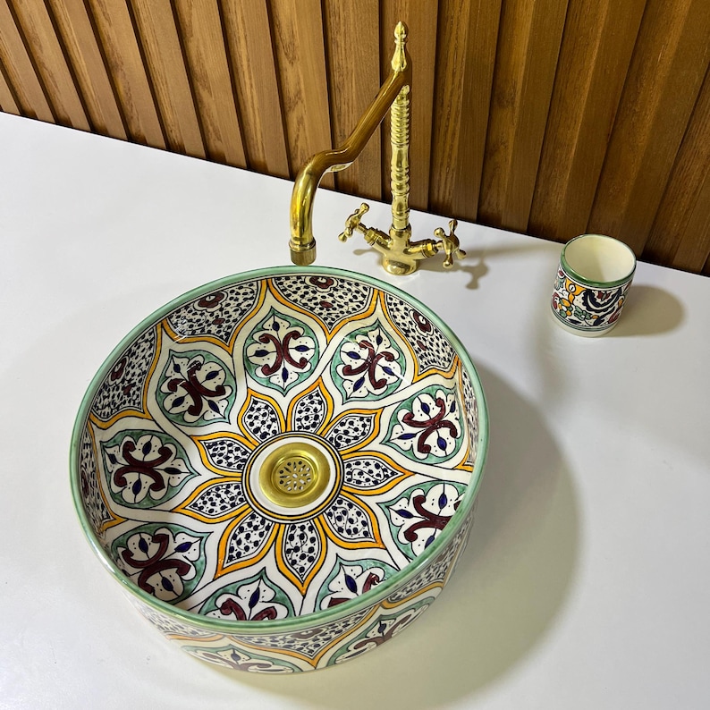 May include: A round ceramic sink with a colorful, intricate floral pattern in shades of green, yellow, red, and blue. The sink has a gold-colored drain and is set against a white background.