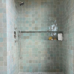 Può includere: Una doccia con piastrelle quadrate azzurre che rivestono le pareti. Il soffione e i rubinetti sono argentati. Un ripiano contiene un asciugamano bianco, una spugna naturale e una spazzola di legno. Il pavimento ha un disegno a piastrelle.