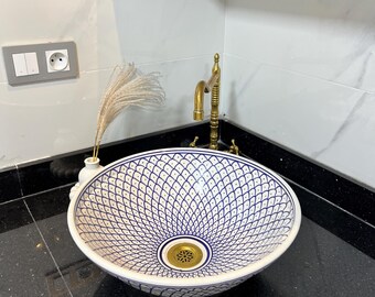 May include: A white ceramic basin with a blue geometric design. The sink features a brass drain and sits on a black countertop. A brass tap is mounted above, and a small vase with dried flowers is placed nearby.
