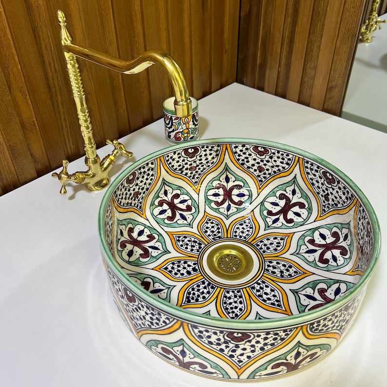 May include: A round ceramic sink with a colorful Moroccan-inspired design. The sink features intricate floral patterns in shades of green, blue, red, and yellow. A gold-toned faucet is mounted above the sink.