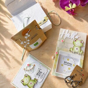 May include: An assortment of handmade greeting cards and gift packaging. The cards feature frog illustrations and the word "Hello". A small box and a brown paper bag are also present, along with a sprig of purple orchids.