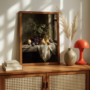 May include: A framed still life painting featuring a wooden table with grapes, plums, pears, and figs. A vase with greenery and a window are visible in the background. The painting is displayed on a wooden cabinet with a lamp and vase.