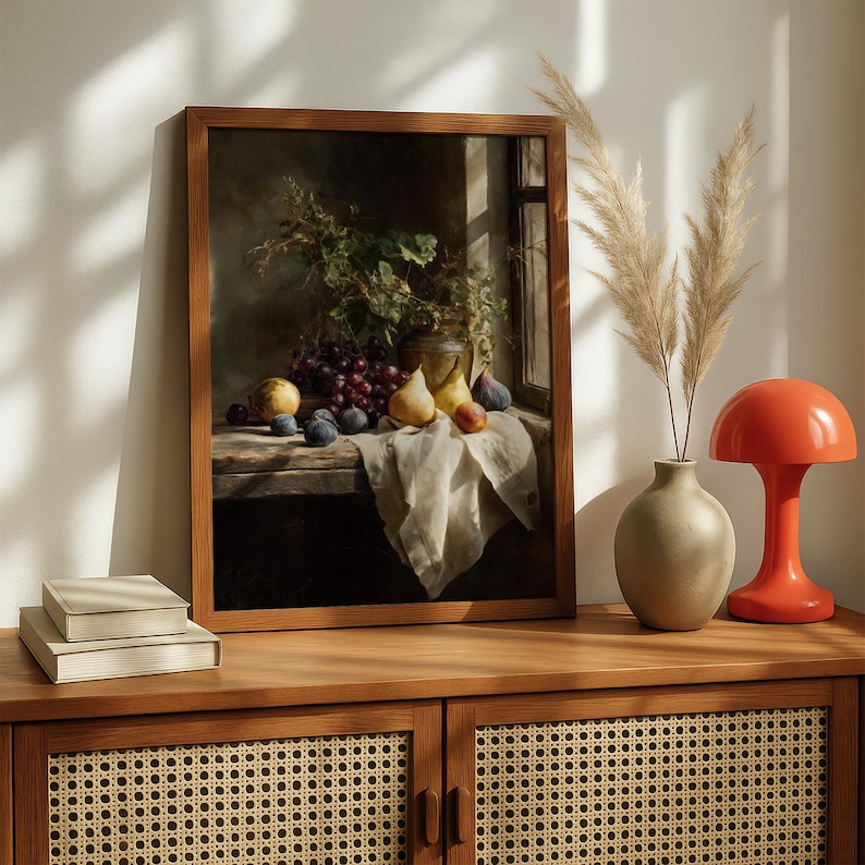 May include: A framed still life painting featuring a wooden table with grapes, plums, pears, and figs. A vase with greenery and a window are visible in the background. The painting is displayed on a wooden cabinet with a lamp and vase.