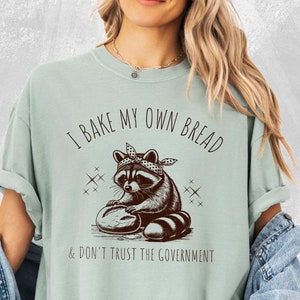 May include: A light green t-shirt with a brown illustration of a raccoon wearing a bandana and holding a loaf of bread. The text "I bake my own bread & don't trust the government" is printed on the shirt.