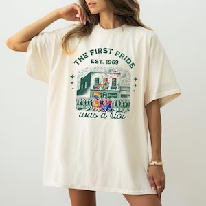May include: Cream-colored t-shirt with a graphic print. The print features a building with a rainbow flag, a crowd of people, and the text "THE FIRST PRIDE EST. 1969 was a riot."