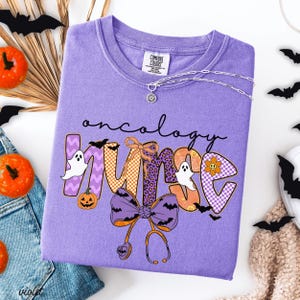 May include: A lilac Comfort Colors sweatshirt with the words "oncology nurse" in a Halloween-themed design. The design includes ghosts, bats, pumpkins, and a stethoscope. A silver necklace with a sun pendant is draped across the shirt.