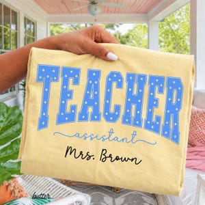 May include: A butter yellow t-shirt featuring the word TEACHER in large blue block letters with white polka dots, the word assistant in blue script, and the name Mrs. Brown printed in black script below the text on the front of the garment.