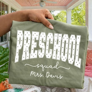 May include: An olive green t-shirt featuring the white text "PRESCHOOL" in a block font with a polka dot pattern, followed by the word "squad" in a cursive script and the name "Mrs. Davis" printed in a white script font below the text.