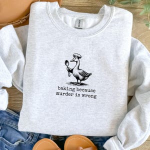 May include: A gray sweatshirt with a black and white illustration of a duck wearing a chef's hat and holding a spoon. The text "baking because murder is wrong" is printed below the illustration.