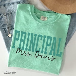 May include: A pale green t-shirt with the word "PRINCIPAL" in teal block letters and "Mrs. Davis" in black script. The shirt is folded on blue jeans and a tan hat.