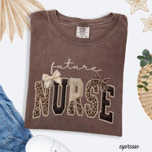 May include: A brown t-shirt with the words "future NURSE" embroidered on the front. The word "NURSE" is in a bold, blocky font with a leopard print pattern. The shirt has a crew neck and short sleeves. The colour is described as espresso.