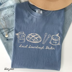 May include: A blue Comfort Colors t-shirt with a graphic of sourdough bread, a jar, utensils, and a flour sack. The text "Local Sourdough Dealer" is printed below the graphic. A tan hat and blue jeans are in the background.