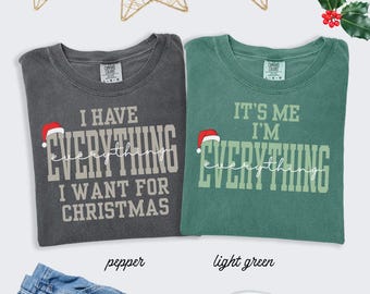 May include: Two Comfort Colors t-shirts, one pepper and one light green, with Christmas-themed text. The pepper shirt says "I HAVE EVERYTHING I WANT FOR CHRISTMAS." The light green shirt says "IT'S ME I'M EVERYTHING." Both shirts have a Santa hat graphic.