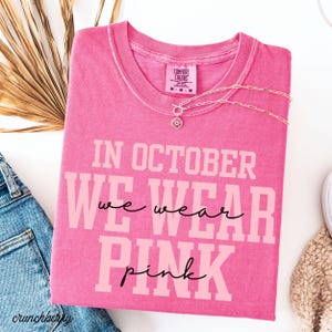 May include: A pink t-shirt with the text "IN OCTOBER WE WEAR PINK" in a bold, light pink font. A silver necklace with a circular pendant is draped over the shirt. The shirt is folded and placed on a white surface.