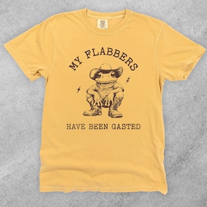 May include: Mustard yellow t-shirt with a graphic of a frog wearing a cowboy hat and boots. The text reads "MY FLABBERS HAVE BEEN GASTED" in a brown, vintage-style font. The frog is squatting, with lightning bolt graphics on either side.